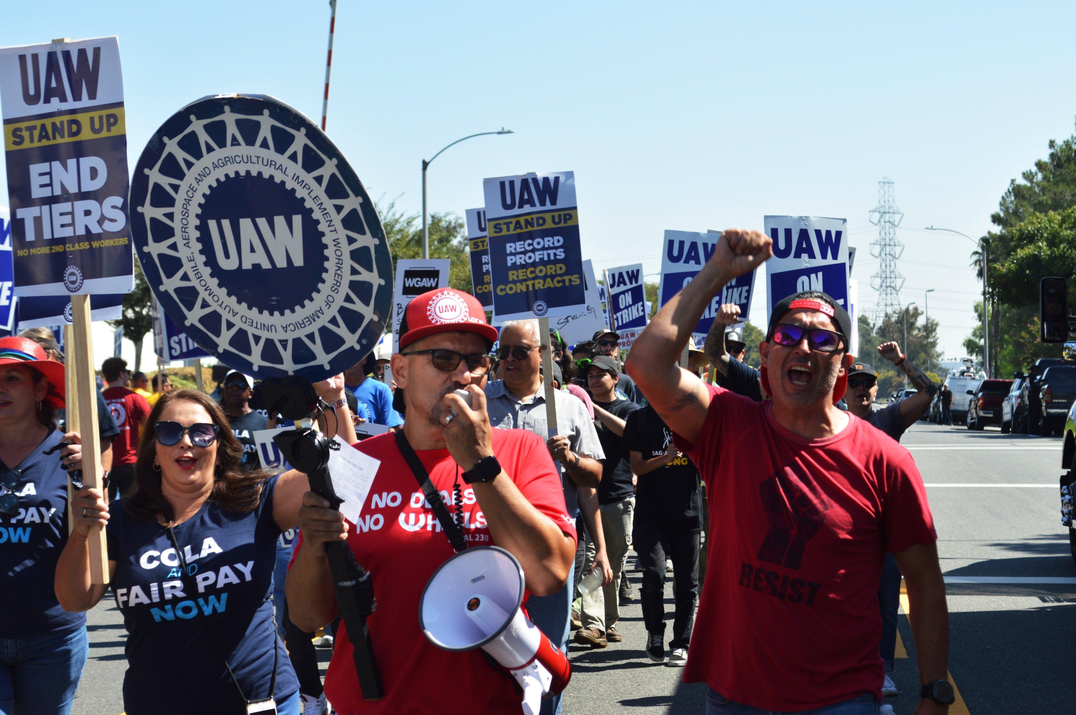 Three UAW 230 members with picket signs chanting near the front of a large march 
