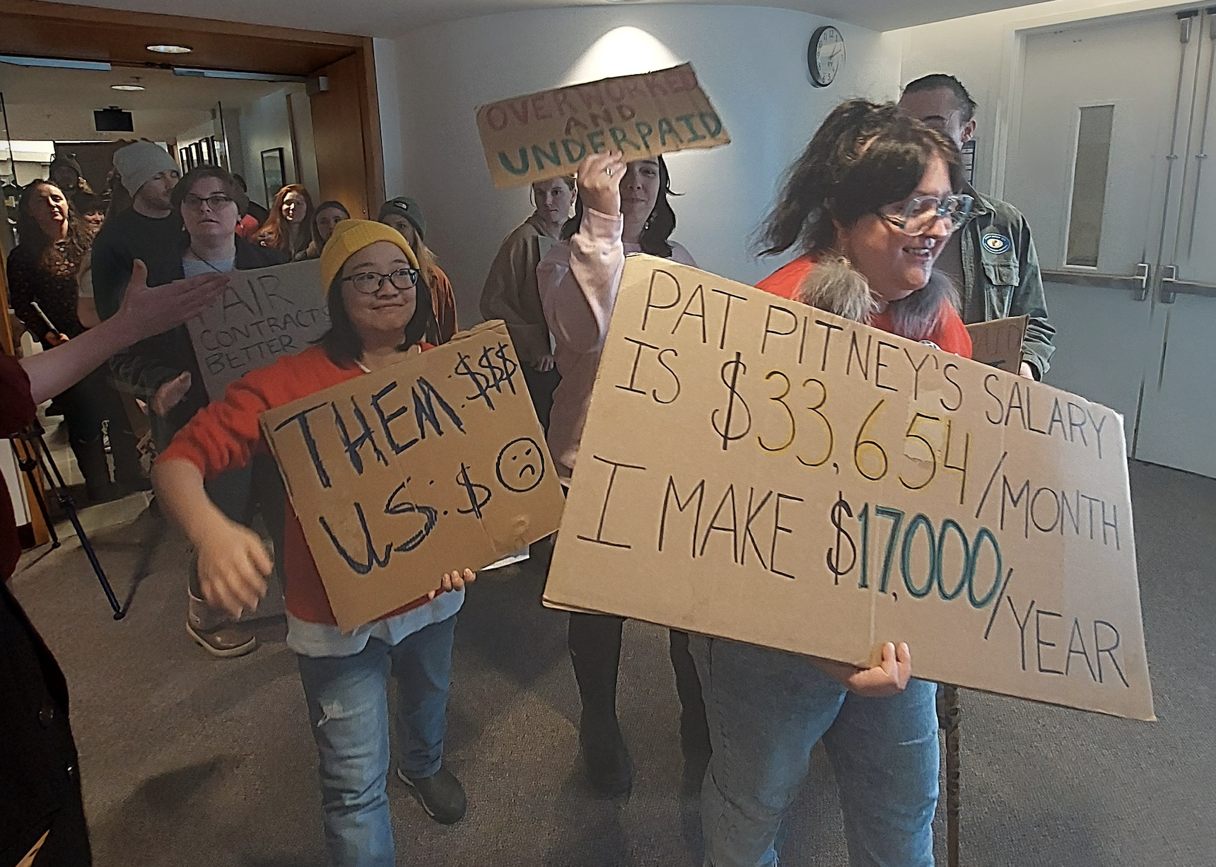 Two AGWA members leading a long line of workers in an indoor hallway. Many workers are holding signs, and one sign in the front says “Pat Pitney’s Salary is $33,654/month. I make $17,000/year.” 