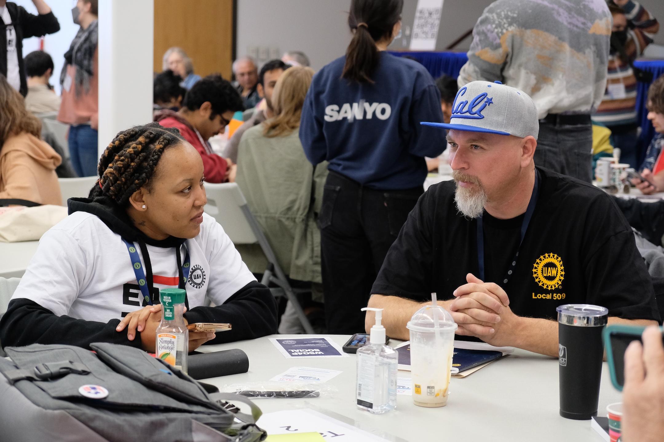 Two UAW 509 members talking at a table, with many others talking at tables in the background