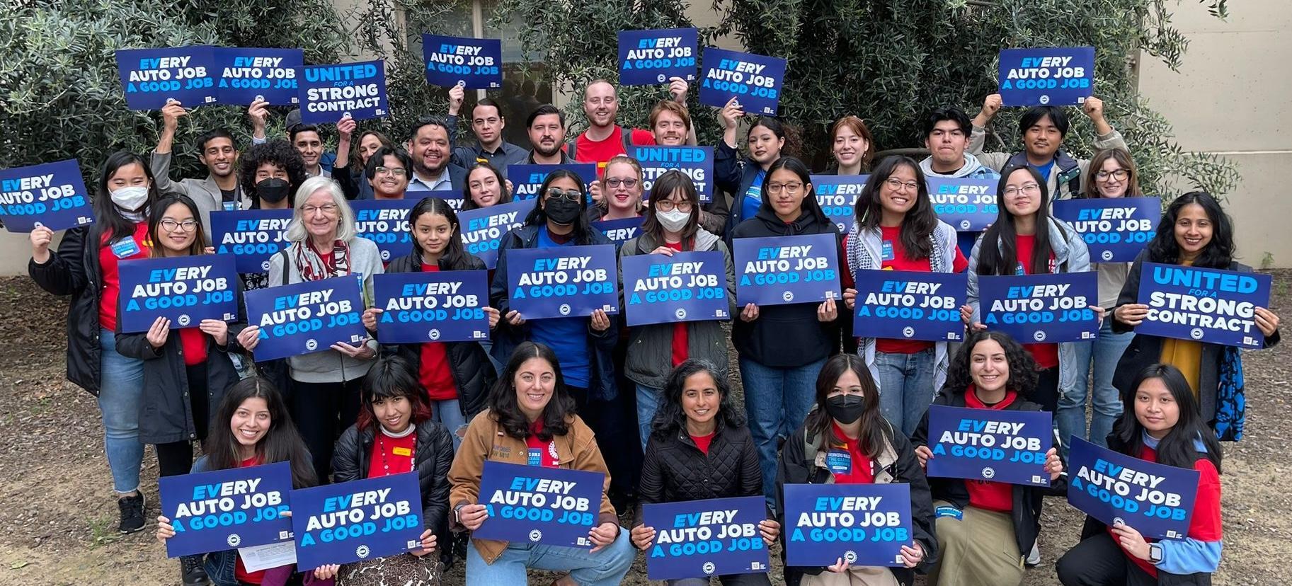A group of about 30 people posed for a photo holding signs reading "EVery auto job a good job"